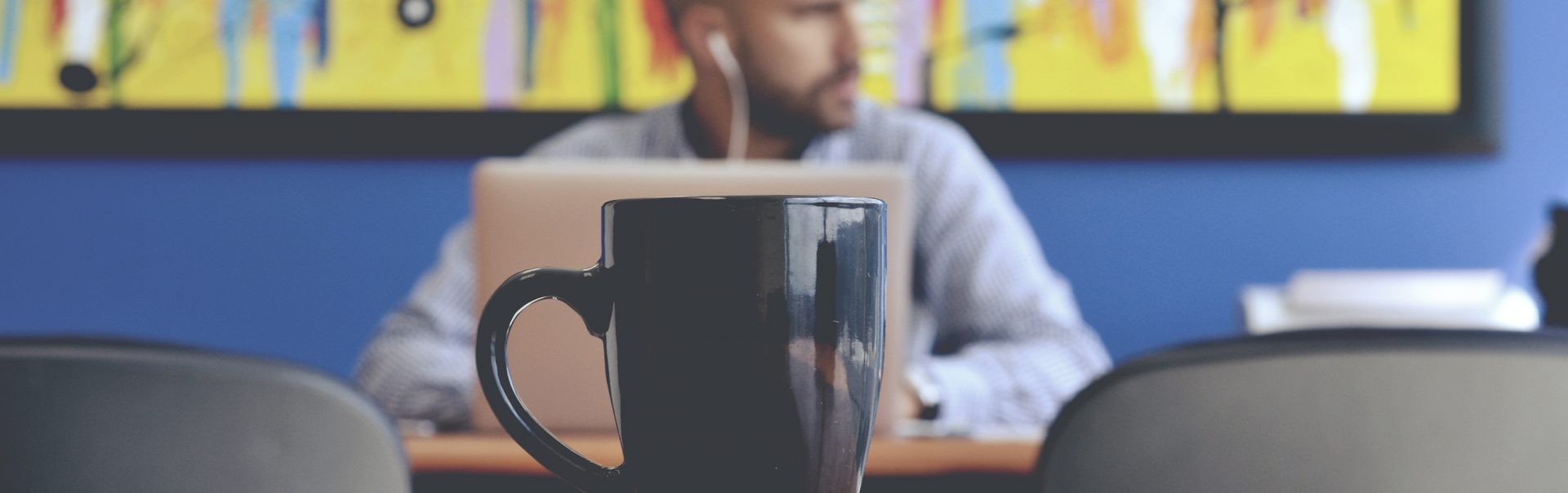 A man works at a cafe in the background. A cup of coffee is in focus in the foreground.