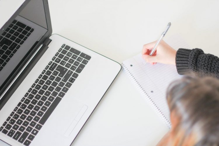 A person, sitting before a computer, takes notes with a pen and paper.