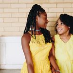 Two teens in yellow dresses laugh in front of a white bench.