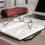 A laptop, notebook, and glasses sit on top of a desk.