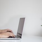 A person types on a computer on a spare white desk with a plant.