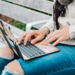 A student works on her computer sitting on a park bench.
