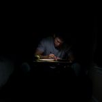 A man sits writing at a desk in the dark.