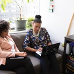 Two women sit on a coach in an office.