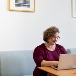 A woman sits down to work at a laptop in a living room setting.