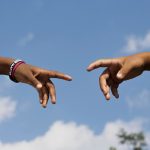 two hands reach out toward one another before the background of a blue sky