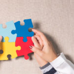 Studio shot, Close-up, Top view of mother's hands holding autistic young child's hands holding colorful jigsaw puzzles (yellow, red, blue, light blue) on beige linen cloth. World autism awareness day support concept.