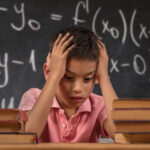 School boy with his head in his hands on a desk behind a chalkboard with math formulas written on it.